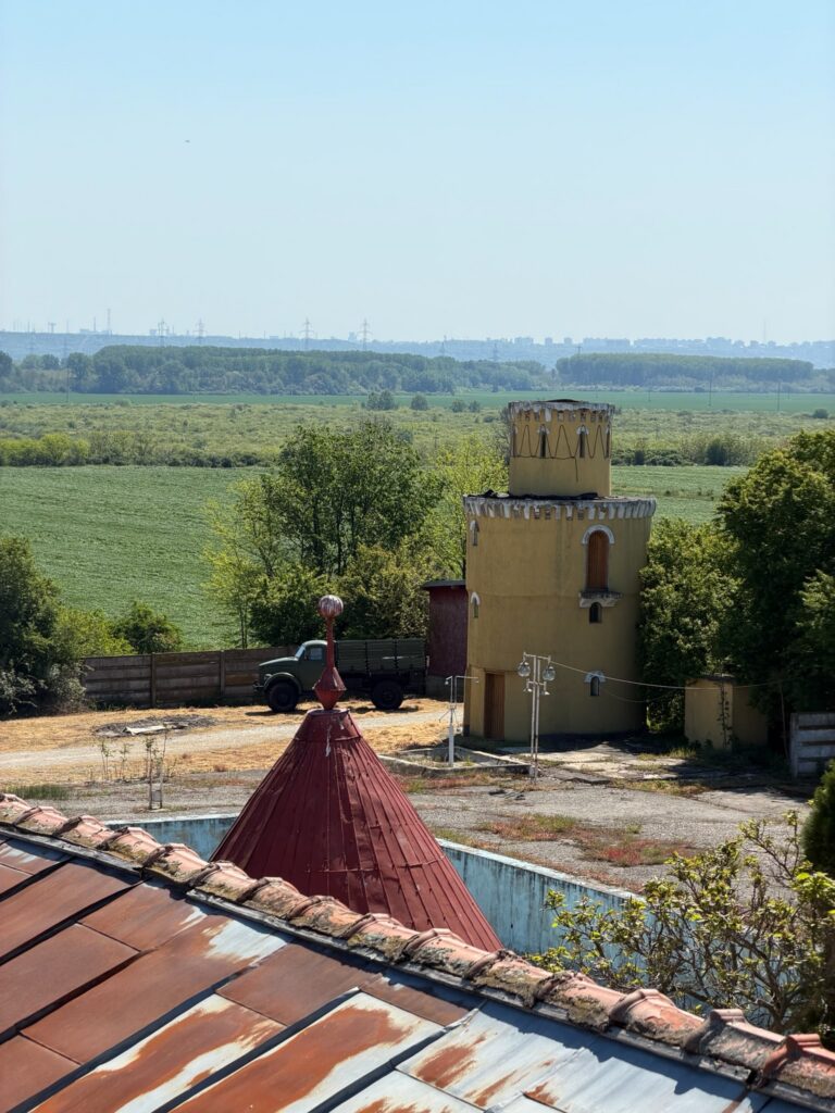 conacul romano plesoiu 8 - Jurnalul Olteniei – Știri din Craiova și Oltenia Yellow cylindrical tower with conical red roof in foreground, rusted metal roof panels, and green fields stretching toward a distant city skyline.