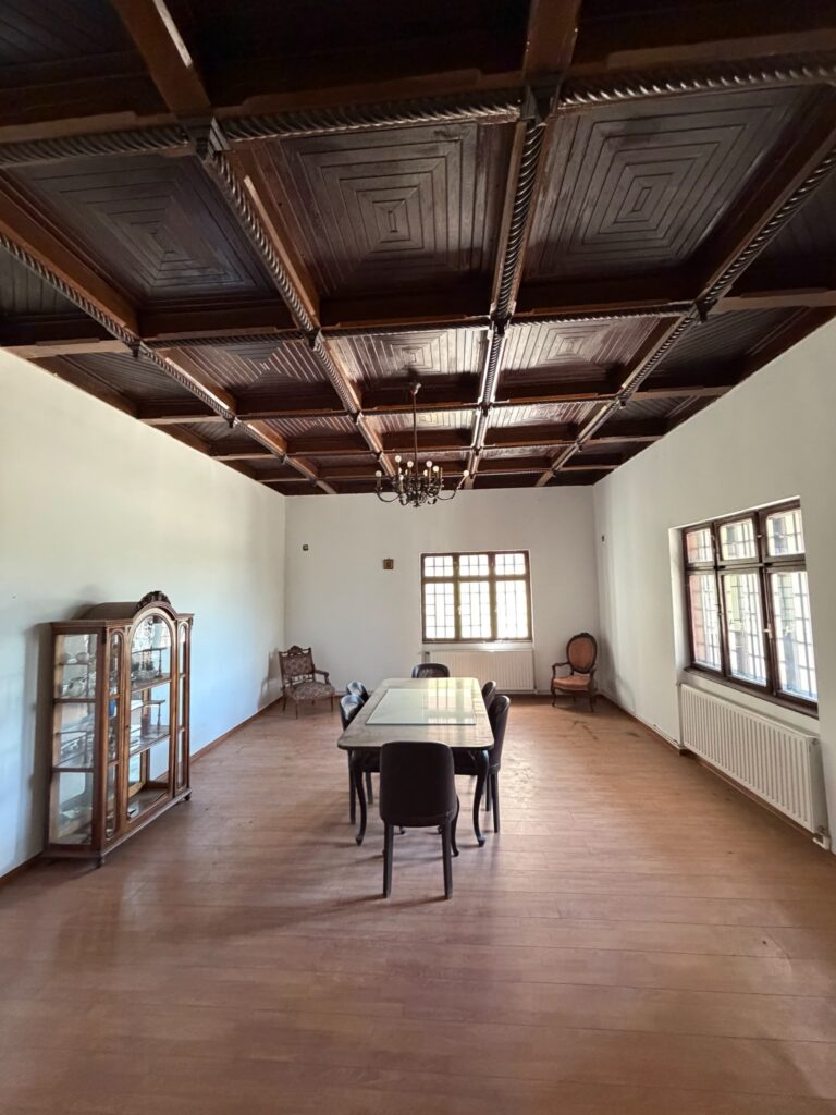 conacul romano plesoiu 9 - Jurnalul Olteniei – Știri din Craiova și Oltenia Dining room with a dark, coffered wooden ceiling, a hanging chandelier, and a long table flanked by chairs; windows along the right wall illuminate the space.