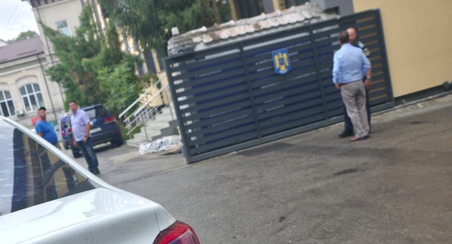 Dark metal gate with a blue-and-yellow emblem; two men in shirts stand nearby, a white car in the foreground, and buildings in the background.