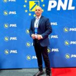 Man in a dark suit posing on a red carpet in front of a blue PNL backdrop with EU stars and a badge visible on his chest.