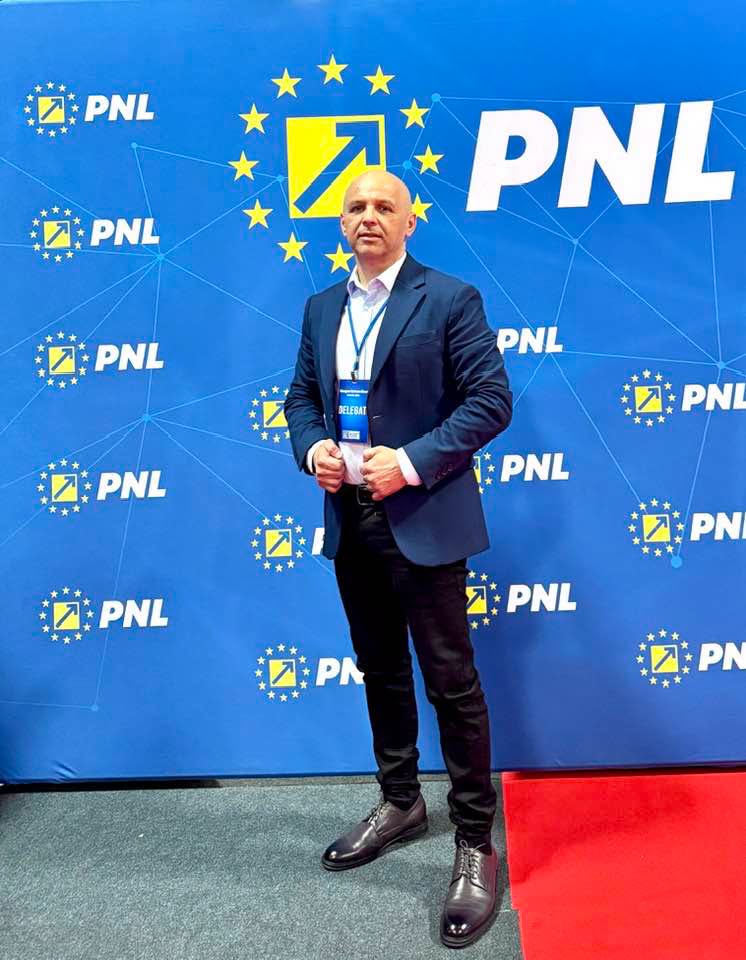 Man in a dark suit posing on a red carpet in front of a blue PNL backdrop with EU stars and a badge visible on his chest.