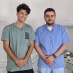 Two men posing in a clinical setting: one in a green T-shirt smiling, the other in blue scrubs with folded hands.