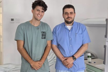 Two men posing in a clinical setting: one in a green T-shirt smiling, the other in blue scrubs with folded hands.