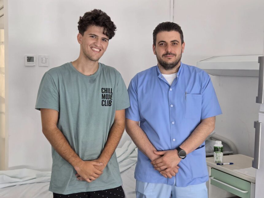 Two men posing in a clinical setting: one in a green T-shirt smiling, the other in blue scrubs with folded hands.