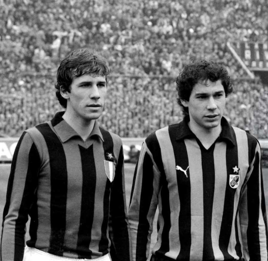 Two soccer players in vertically striped uniforms stand side by side on a field with a crowded stadium in the background, focused on something ahead.
