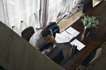 Overhead view of a person at a dark wooden desk, laptop open, papers spread out, with a plant nearby.