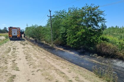 Fire truck with firefighters on a dirt road beside a scorched ditch and burned vegetation under a clear blue sky.