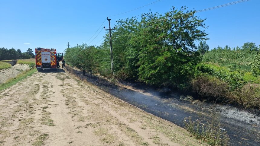 Fire truck with firefighters on a dirt road beside a scorched ditch and burned vegetation under a clear blue sky.