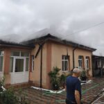Beige two-story house with orange trim; steam or smoke rising from the roof while hoses lie on the tiled yard and a man stands in the foreground.
