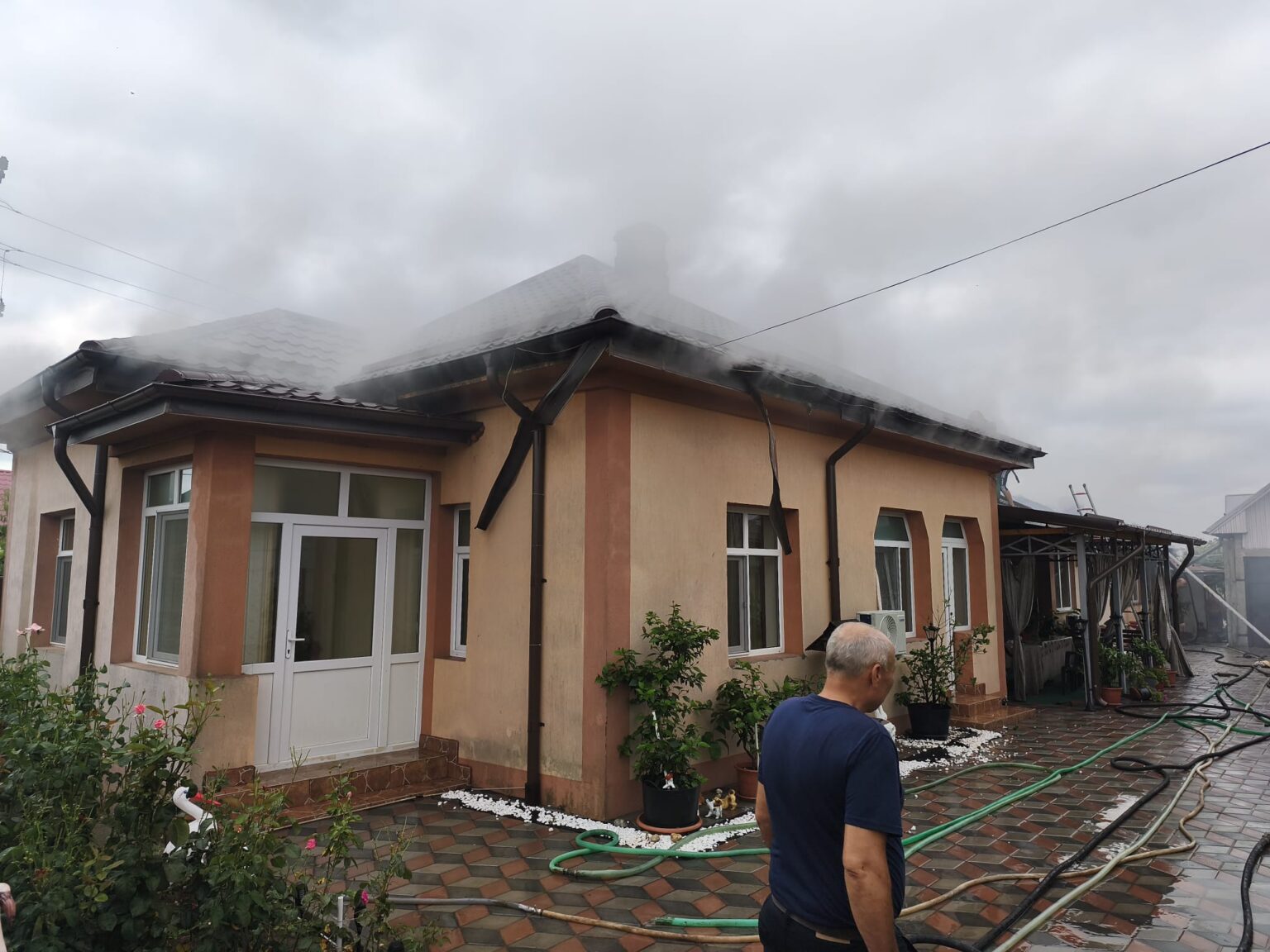 incendiu piatra Olt 2 - Jurnalul Olteniei – Știri din Craiova și Oltenia Beige two-story house with orange trim; steam or smoke rising from the roof while hoses lie on the tiled yard and a man stands in the foreground.