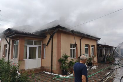 Beige two-story house with orange trim; steam or smoke rising from the roof while hoses lie on the tiled yard and a man stands in the foreground.