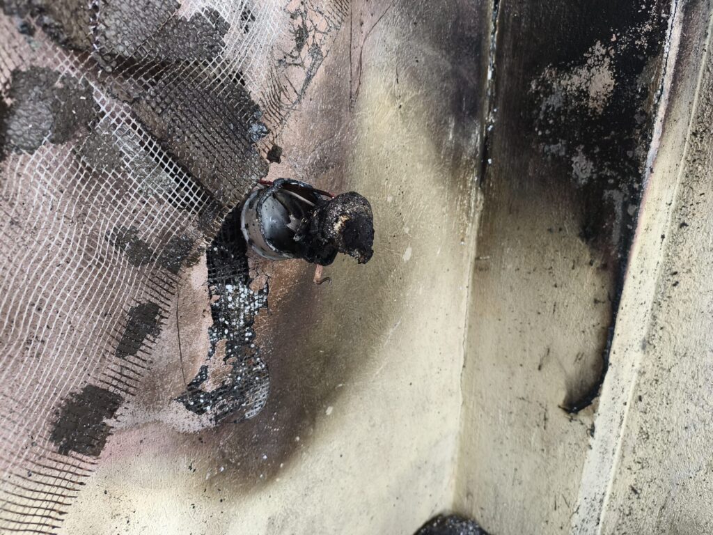 incendiu piatra Olt 3 - Jurnalul Olteniei – Știri din Craiova și Oltenia Close-up of a charred, soot-covered wall with cracked plaster, a grid-like metal mesh, and a damaged circular metal component at the center.