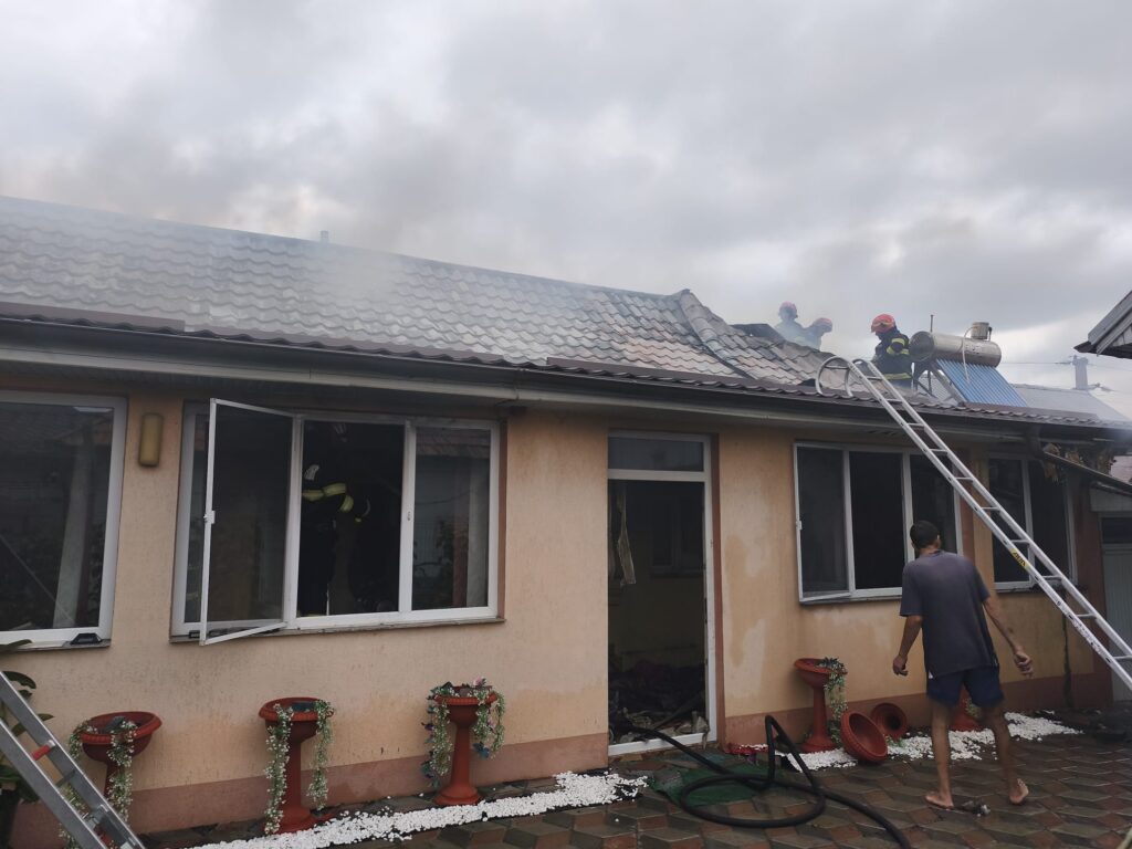 incendiu piatra Olt 4 - Jurnalul Olteniei – Știri din Craiova și Oltenia Firefighters on a smoke-filled roof of a beige house, using a ladder and hoses as debris and smoke surround the scene.