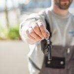 Person in gray shirt and overalls holds out car keys toward the camera (handing over keys).