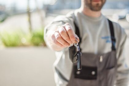 Person in gray shirt and overalls holds out car keys toward the camera (handing over keys).