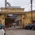 Entrance to a yellow industrial building with a gate, Romanian flags, and signs reading Uzina Mecánica Sadu-Gorj (Romania)