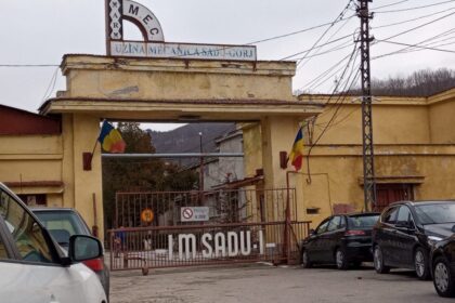 Entrance to a yellow industrial building with a gate, Romanian flags, and signs reading Uzina Mecánica Sadu-Gorj (Romania)
