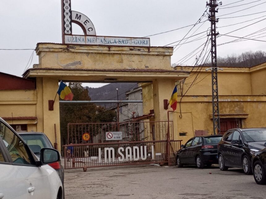 Entrance to a yellow industrial building with a gate, Romanian flags, and signs reading Uzina Mecánica Sadu-Gorj (Romania)