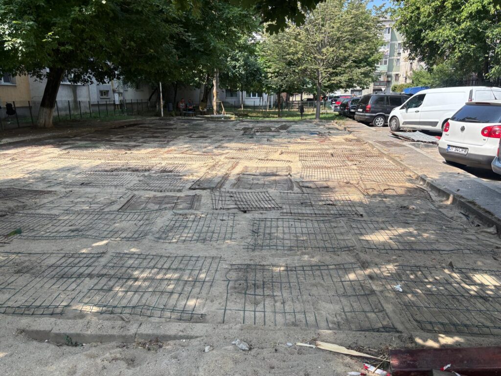 Open parking area under repair with exposed steel rebar grid prepared for new pavement, cars parked along the curb and trees in the background.