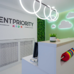 Reception area of a pediatric dental clinic with a 'DENTPRIORITY KIDS' sign, white desk, and cloud-themed green wall/panel decor.