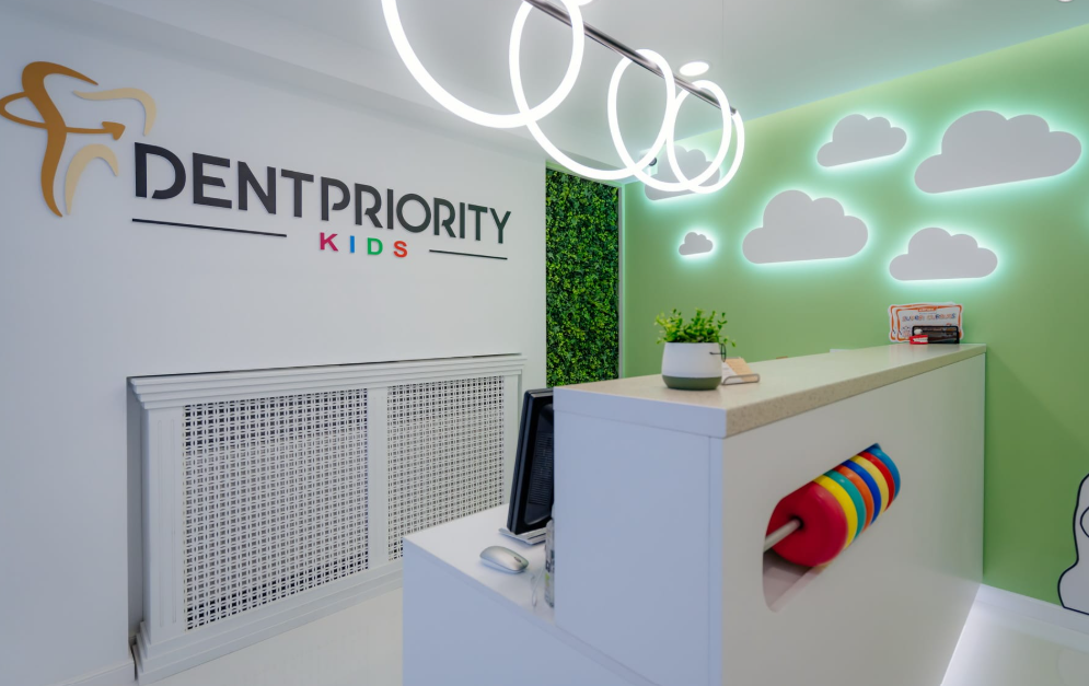Reception area of a pediatric dental clinic with a 'DENTPRIORITY KIDS' sign, white desk, and cloud-themed green wall/panel decor.