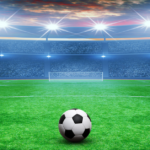 Soccer ball on a green field facing a large stadium under bright floodlights.