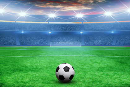Soccer ball on a green field facing a large stadium under bright floodlights.