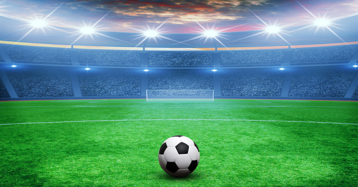 Soccer ball on a green field facing a large stadium under bright floodlights.