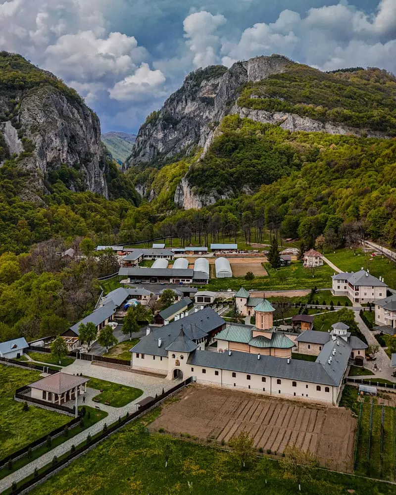 Aerial view of a small mountain valley village with white buildings, a central church, and surrounding green hills and towering cliffs in the background.