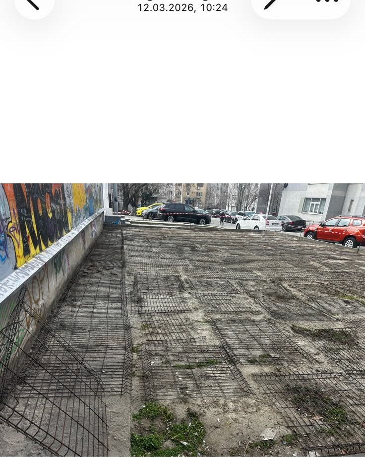 Urban outdoor area with exposed steel rebar grids on the ground, graffiti-covered wall on the left, and several parked cars in the background.