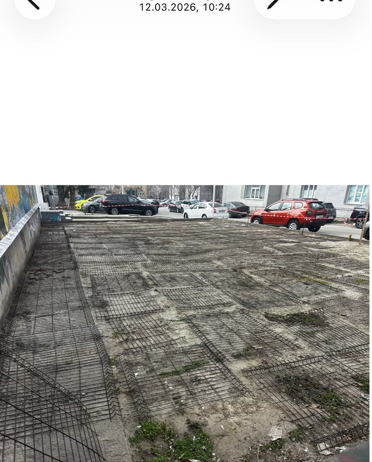 Open parking lot with exposed metal rebar grid preparing for concrete, surrounding vehicles in background.