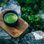 Cup of frothy green matcha tea in a dark bowl resting on a wooden board outdoors among rocks and greenery.