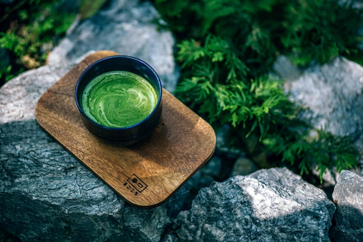 matcha - Jurnalul Olteniei – Știri din Craiova și Oltenia Cup of frothy green matcha tea in a dark bowl resting on a wooden board outdoors among rocks and greenery.