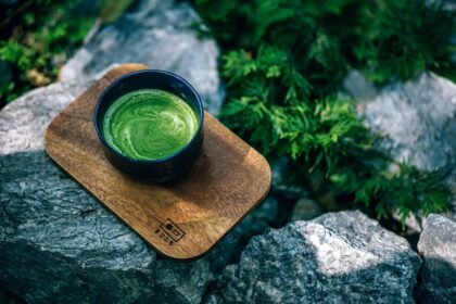 Cup of frothy green matcha tea in a dark bowl resting on a wooden board outdoors among rocks and greenery.