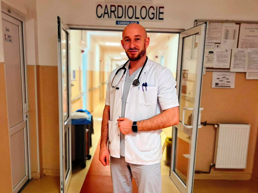 medic cardiolg nou - Jurnalul Olteniei – Știri din Craiova și Oltenia Male doctor in a white coat with a stethoscope stands in a hospital corridor, looking at the camera.