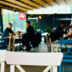 Blurred cafe interior with patrons seated at tables, colorful chairs, and string lights along a glass wall.