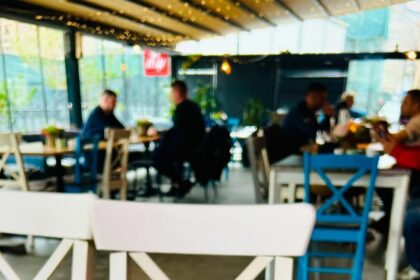 Blurred cafe interior with patrons seated at tables, colorful chairs, and string lights along a glass wall.