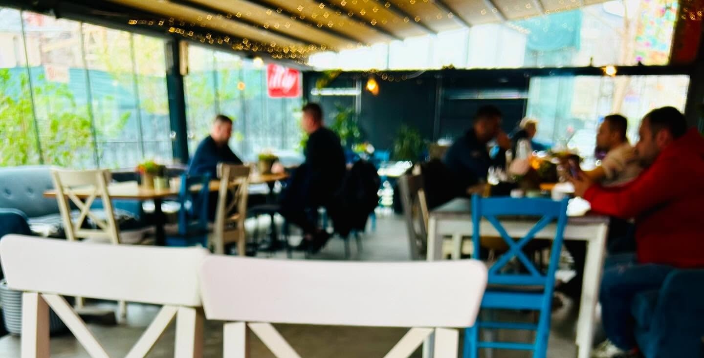 Blurred cafe interior with patrons seated at tables, colorful chairs, and string lights along a glass wall.