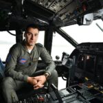 Military pilot in cockpit, seated at the controls, wearing an olive flight suit with patches (Airshow patch visible).