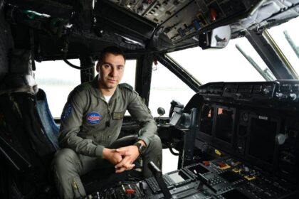 Military pilot in cockpit, seated at the controls, wearing an olive flight suit with patches (Airshow patch visible).