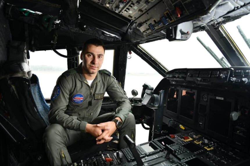 Military pilot in cockpit, seated at the controls, wearing an olive flight suit with patches (Airshow patch visible).