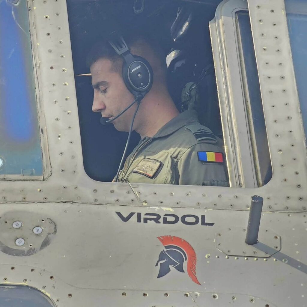 mihai virdol 758830839_1341871621458191_8162634172677243817_n - Jurnalul Olteniei – Știri din Craiova și Oltenia Pilot in headset seated in a cockpit window of a military helicopter, Romanian flag patch on the uniform sleeve, looking downward.