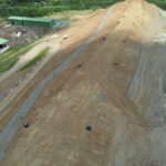 Aerial view of a large dirt ramp at a construction site with green storage buildings to the left and a curved access road lined with red and white cones.
