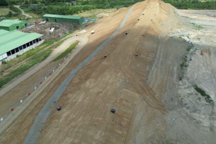 Aerial view of a large dirt ramp at a construction site with green storage buildings to the left and a curved access road lined with red and white cones.