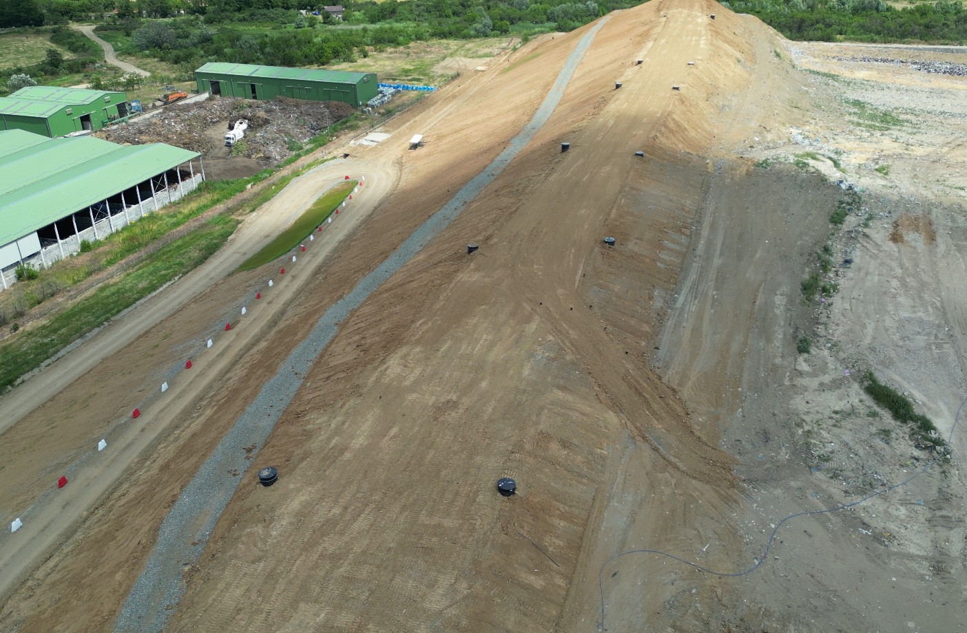 Aerial view of a large dirt ramp at a construction site with green storage buildings to the left and a curved access road lined with red and white cones.
