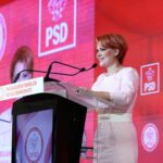 Woman in a pale pink dress delivering a speech at a podium with PSD banners in a red-themed backdrop.