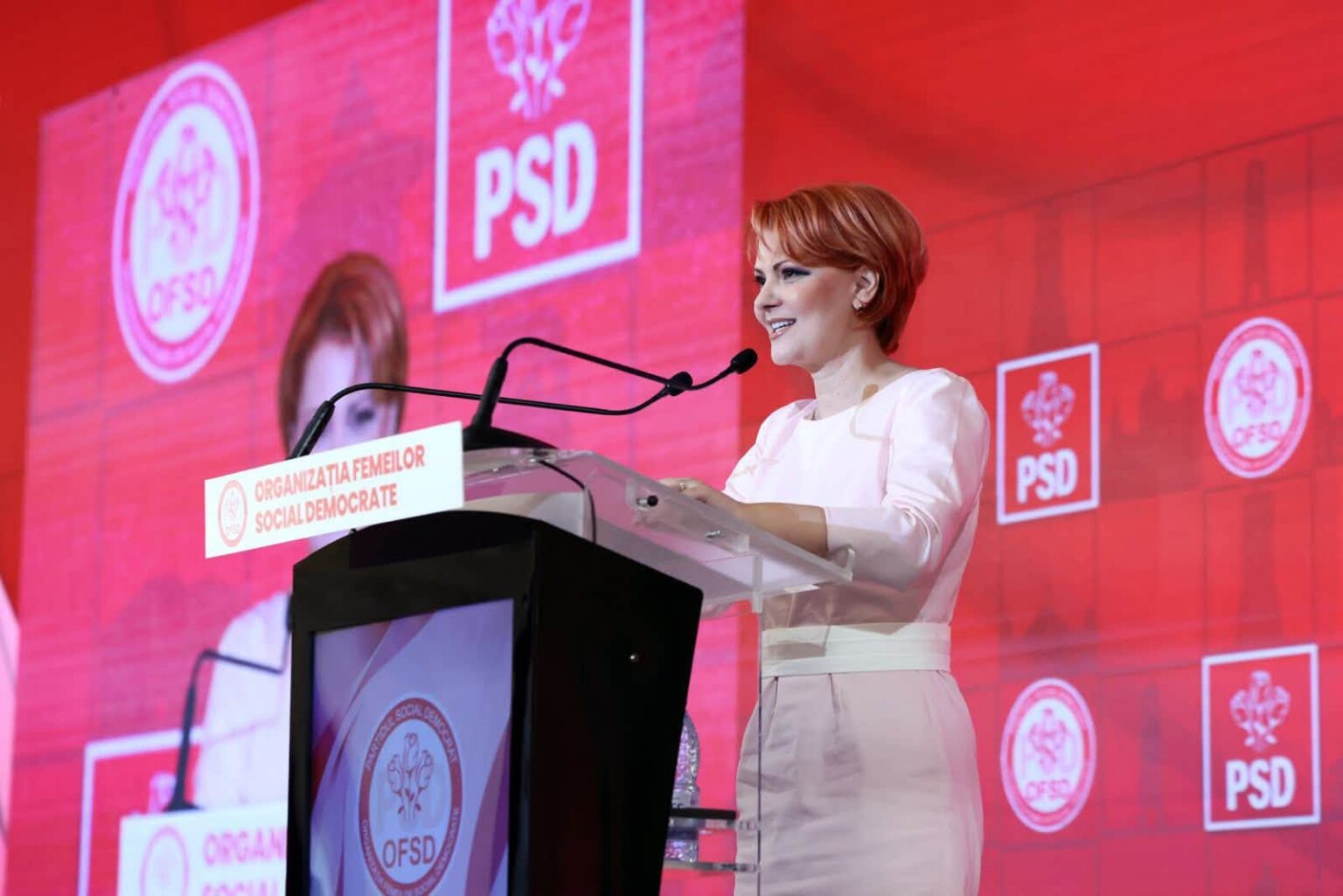 Woman in a pale pink dress delivering a speech at a podium with PSD banners in a red-themed backdrop.