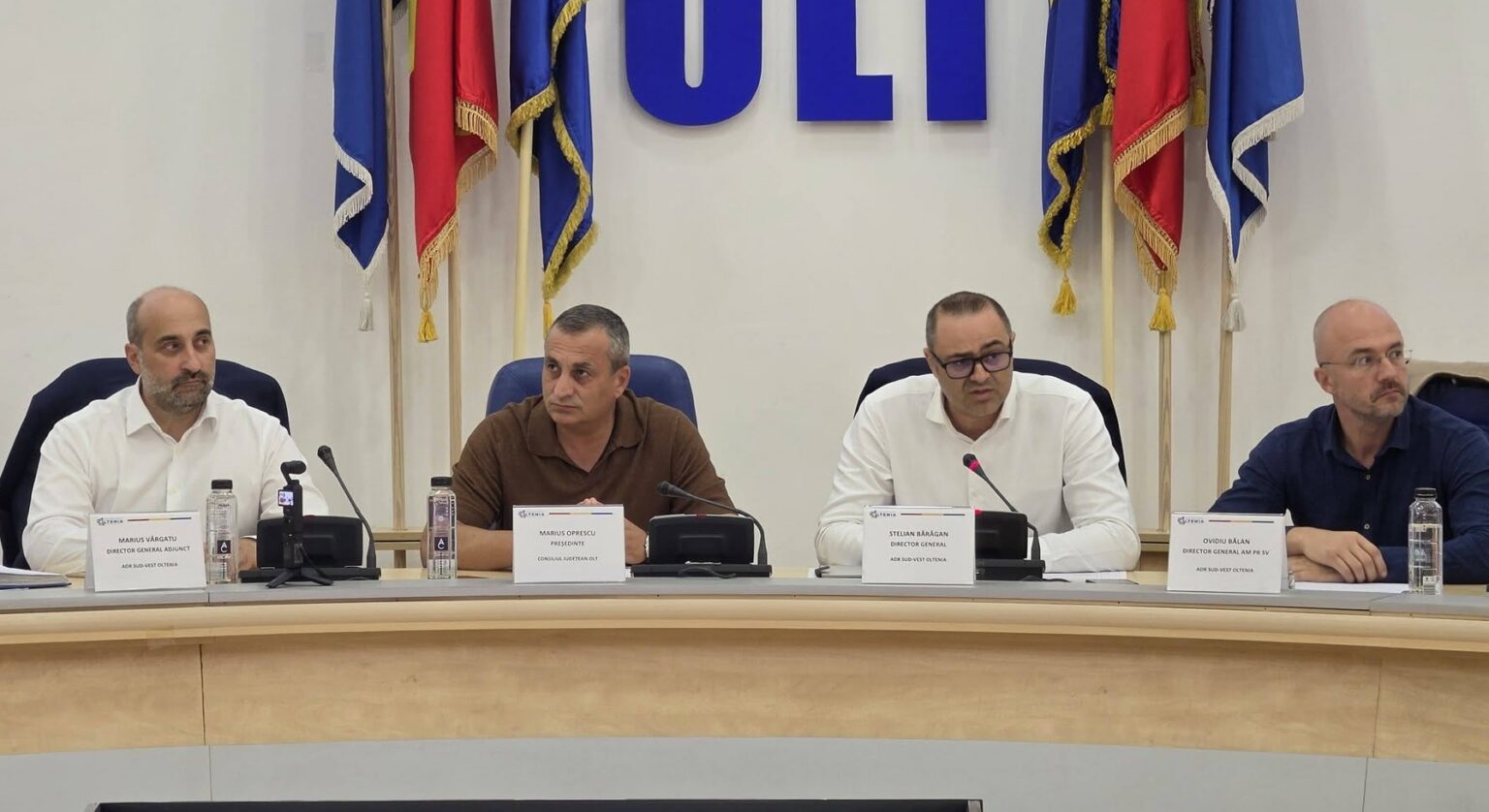 Four men seated at a conference table with microphones and nameplates, flags behind them in a formal panel setting.