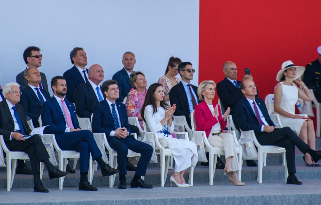 Group of formally dressed officials seated in white chairs on steps at an outdoor ceremony, some clapping, with a blue sky and red backdrop behind them.