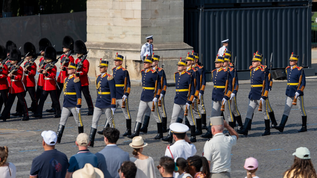 Rows of soldiers marching in blue uniforms with gold trim and yellow belts, flanked by red-coated guards with tall black bearskin hats, as spectators watch.